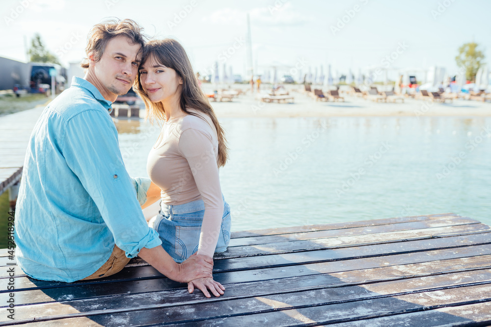 Couple of two, man and woman, sitting together on pier of wooden bridge in the sea or lake and hugging. Leisure, love relationships and people concept.