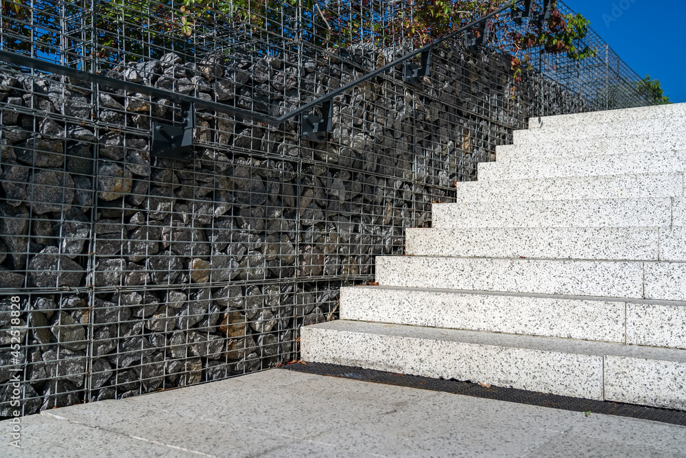 Granite staircase with fence made of stones in metal grid. Modern concrete steps with gabion fence. Modern broad stairs with stone wall and grey metallic handrail