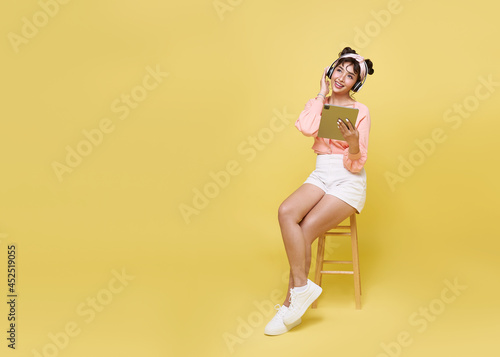 Happy beautiful Asian teen woman sitting on chair and hand holding tablet with wearing wireless headphones listening to music on yellow copy space background.