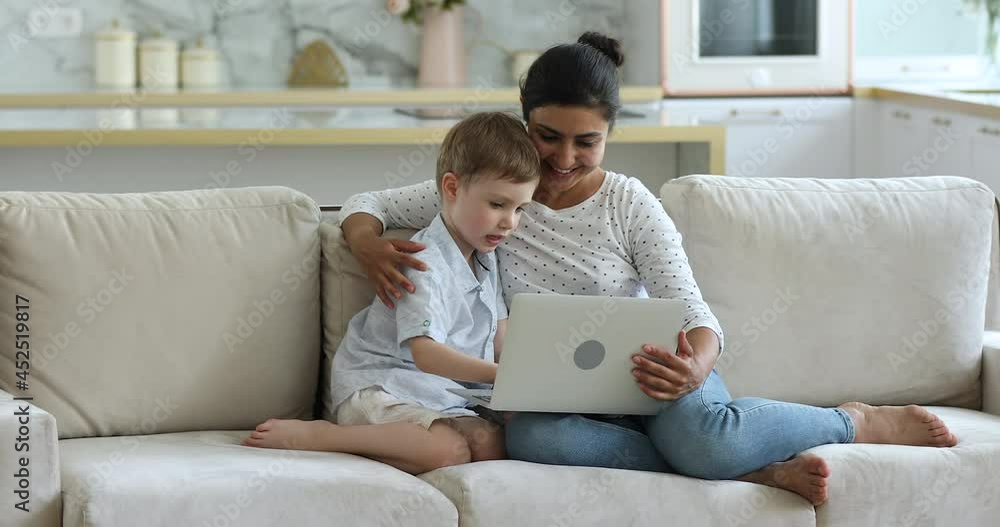 Young Indian mother Caucasian adopted son, nanny and little preschooler 5s boy sit together on couch at home with laptop. Develop teach child use wireless device, play educational game on-line concept Young Indian mother Caucasian adopted son, nanny and little preschooler 5s boy sit together on couch at home with laptop. Develop teach child use wireless device, play educational game on-line concept