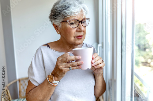 Thoughtful old woman in beige blouse drinking mug of coffee and looking through window at home. Beautiful senior woman at home standing at the window in her living room holding a cup of coffee or tea.