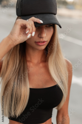 Cool beautiful young blonde caucasian woman in a fashion black top wears a fashionable black cap and walks in the city