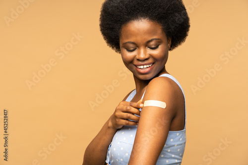 Leinwand Poster Vaccinated african woman showing arm with adhesive bandage after vaccine injection for covid prevention with happy smile isolated over beige studio wall