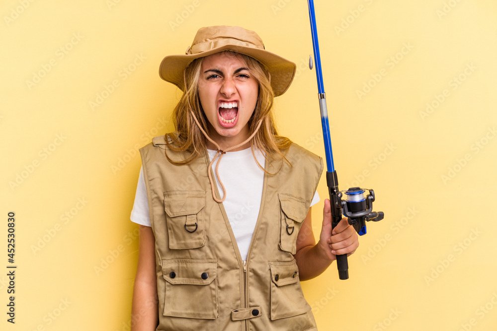 Young caucasian blonde fisherwoman holding a rod isolated on yellow background screaming very angry and aggressive. Young caucasian blonde fisherwoman holding a rod isolated on yellow background screaming very angry and aggressive.