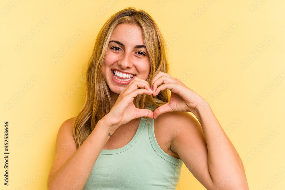 Young caucasian blonde woman isolated on yellow background smiling and showing a heart shape with hands. Young caucasian blonde woman isolated on yellow background smiling and showing a heart shape with hands.
