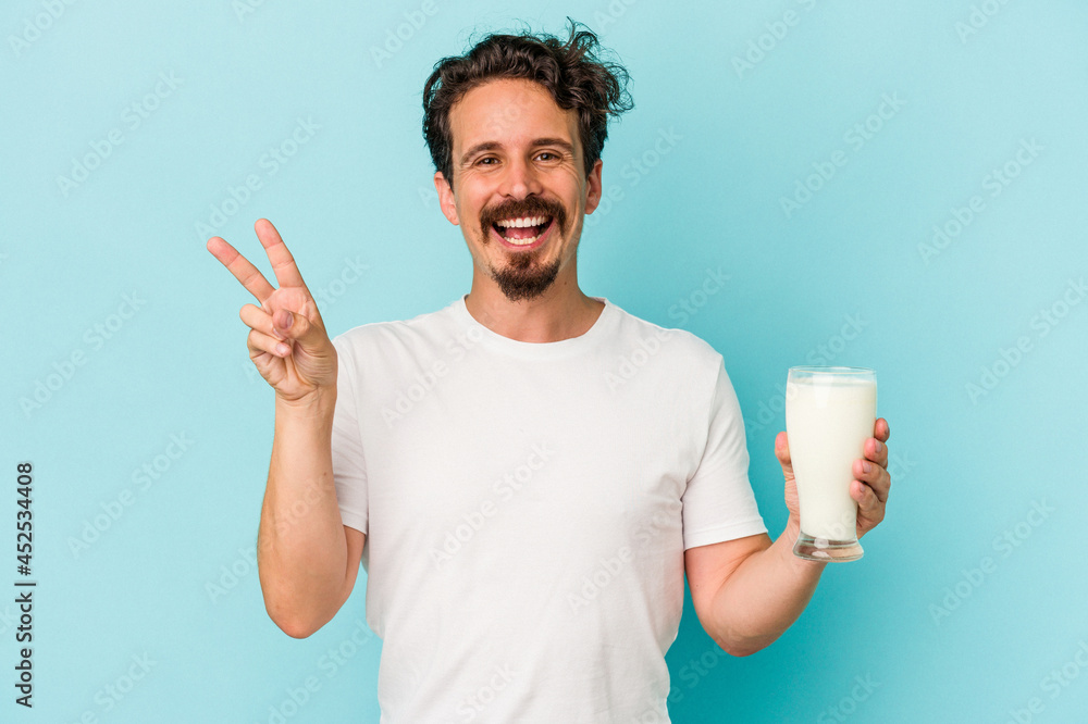 Young caucasian man holding a glass of milk isolated on blue background joyful and carefree showing a peace symbol with fingers. Young caucasian man holding a glass of milk isolated on blue background joyful and carefree showing a peace symbol with fingers.