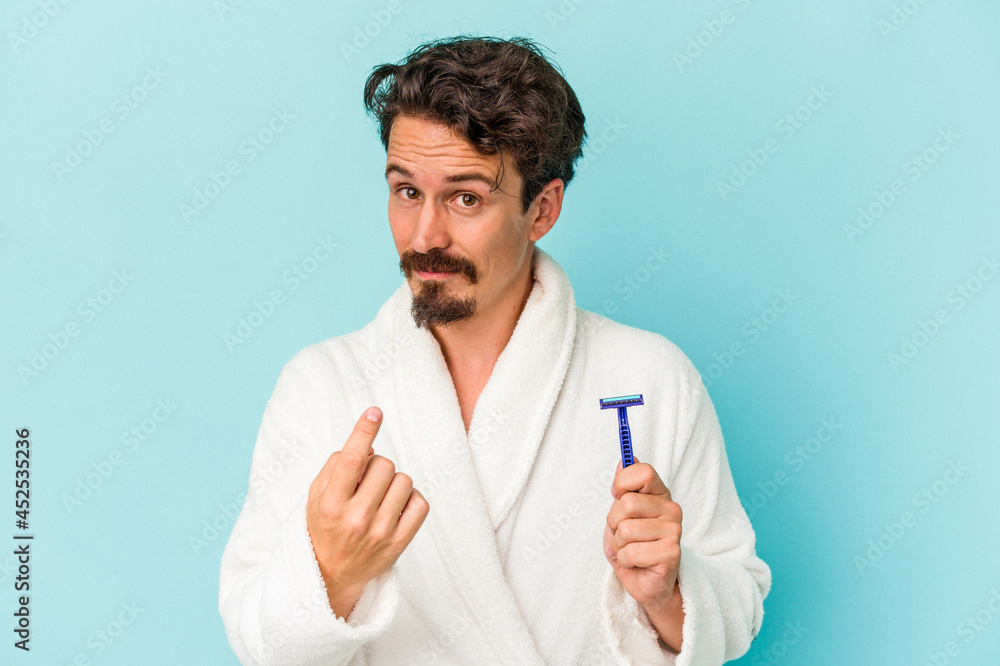 Young caucasian man holding a razor blade isolated on blue background pointing with finger at you as if inviting come closer. Young caucasian man holding a razor blade isolated on blue background pointing with finger at you as if inviting come closer.