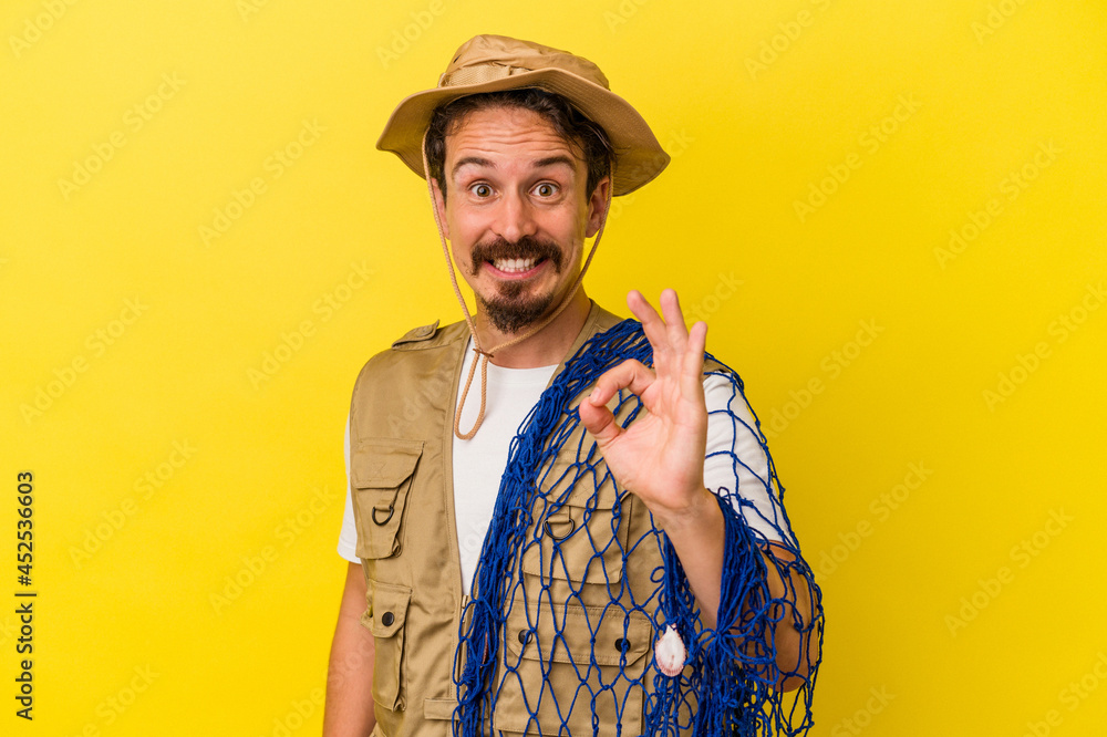 Young caucasian fisherman holding net isolated on yellow background cheerful and confident showing ok gesture. Young caucasian fisherman holding net isolated on yellow background cheerful and confident showing ok gesture.