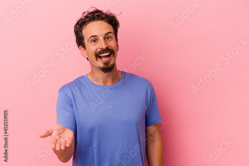 Young caucasian man isolated on pink background stretching hand at camera in greeting gesture. © Asier