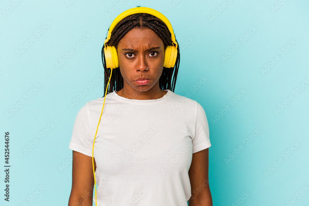 Fototapeta premium Young African American woman listening to music isolated on blue background shrugs shoulders and open eyes confused.