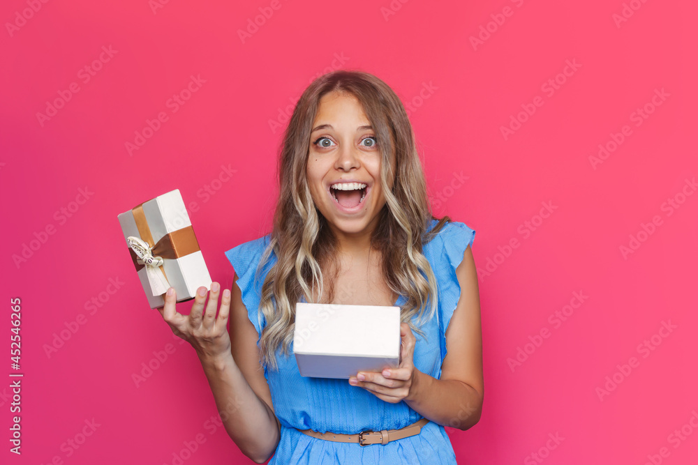 A young charming amazed excited blonde woman with wavy hair in a blue dress is happy with the gift she received isolated on a color pink background. A satisfied girl got a pleasant surprise A young charming amazed excited blonde woman with wavy hair in a blue dress is happy with the gift she received isolated on a color pink background. A satisfied girl got a pleasant surprise