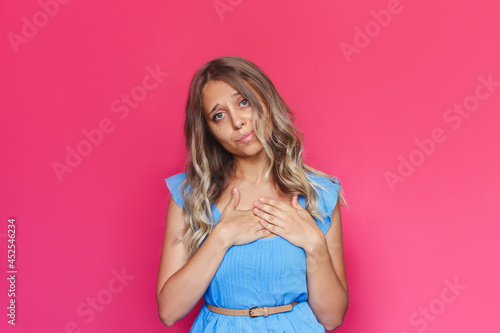 A young charming blonde woman with wavy hair in a blue dress holds her hands on her chest sincerely apologize or regret isolated on a bright color pink background. Gratitude concept