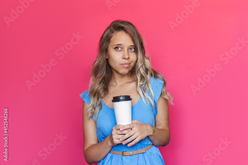 A young charming blonde woman with wavy hair in a blue dress holds a white paper cup of coffee or tea with copy space looking away isolated on a bright color pink background. Mockup for text or logo