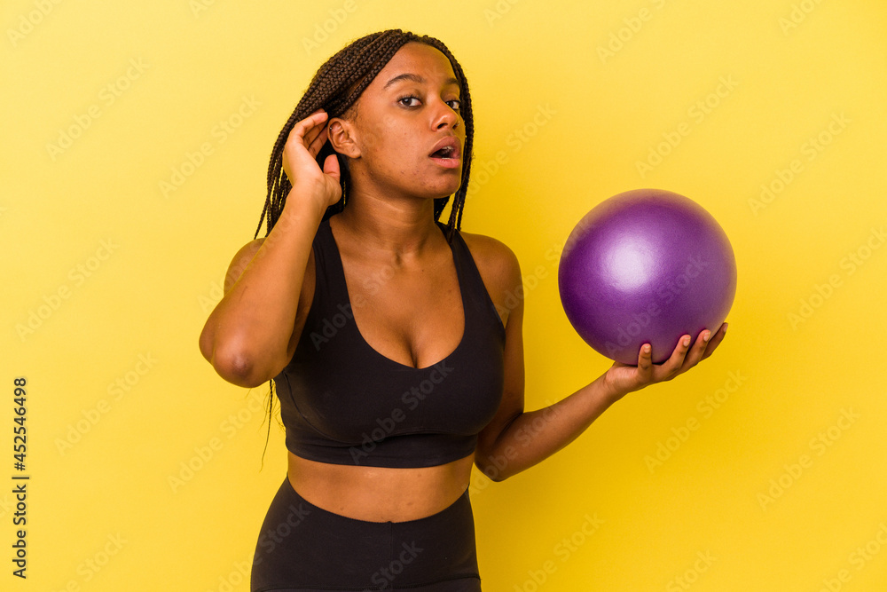 Young african american woman holding a pilates ball isolated on yellow background trying to listening a gossip. Young african american woman holding a pilates ball isolated on yellow background trying to listening a gossip.