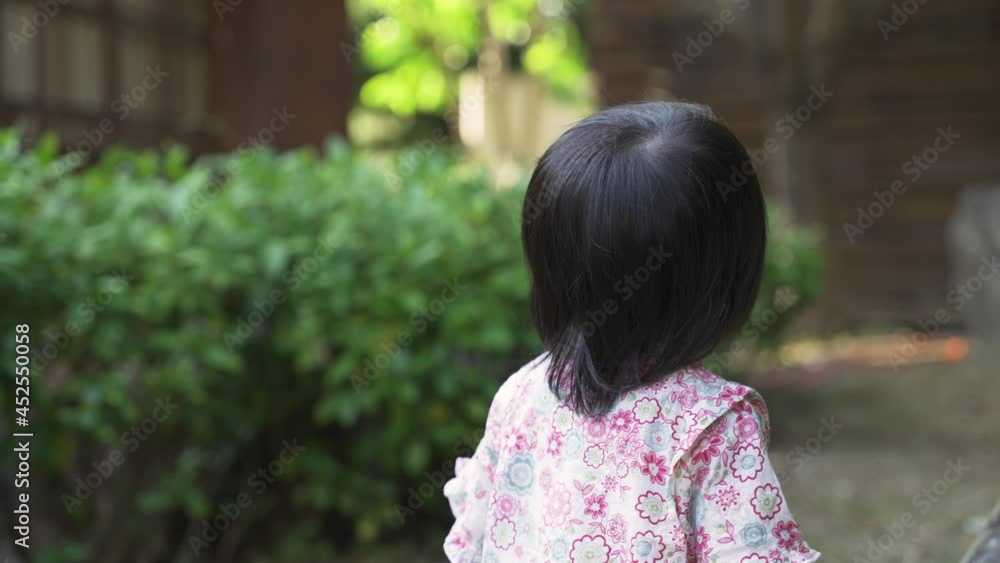 curious Asian toddler girl wearing kimono is turning back on camera and looking into distance while playing by a large stone in the yard on a sunny day curious Asian toddler girl wearing kimono is turning back on camera and looking into distance while playing by a large stone in the yard on a sunny day
