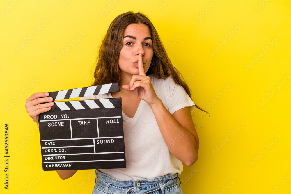 Young caucasian woman holding clapperboard isolated on yellow background  keeping a secret or asking for silence.