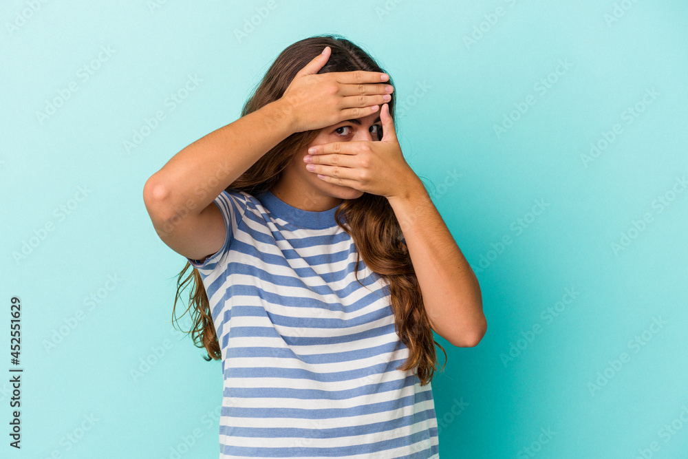 Young caucasian woman isolated on blue background blink at the camera through fingers, embarrassed covering face. Young caucasian woman isolated on blue background blink at the camera through fingers, embarrassed covering face.