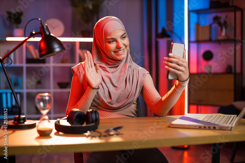 Attractive arabian woman using modern cell phone for taking selfie. Positive young female in hijab sitting at desk with wireless laptop and having break during work at evening time.