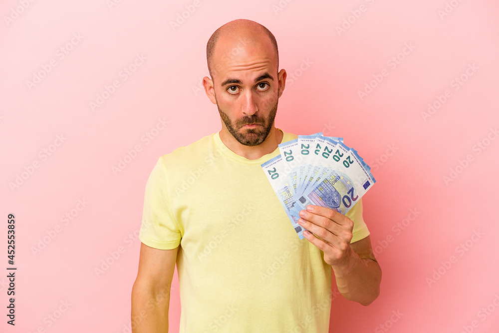 Young caucasian bald man holding bills isolated on pink background shrugs shoulders and open eyes confused. Young caucasian bald man holding bills isolated on pink background shrugs shoulders and open eyes confused.