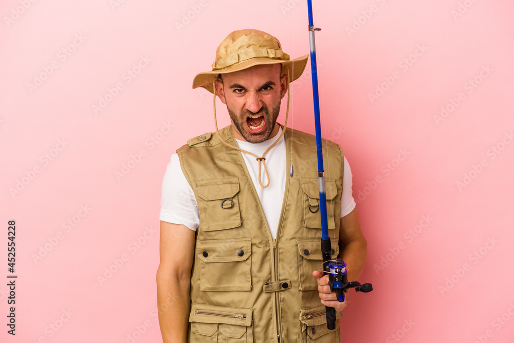 Young bald fisherman holding a rod isolated on pink background screaming very angry and aggressive. Young bald fisherman holding a rod isolated on pink background screaming very angry and aggressive.