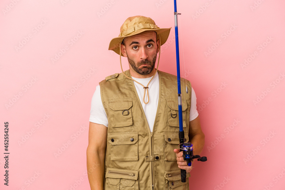 Young bald fisherman holding a rod isolated on pink background shrugs shoulders and open eyes confused. Young bald fisherman holding a rod isolated on pink background shrugs shoulders and open eyes confused.