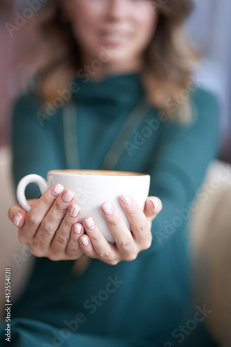 Blurred background. A beautiful woman with a beautiful manicure holds a cup of coffee and stretches it forward. She looks directly at the camera with a smile. High quality photo