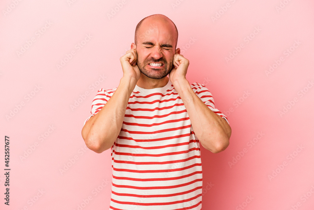 Fototapeta premium Young caucasian bald man isolated on pink background covering ears with hands.