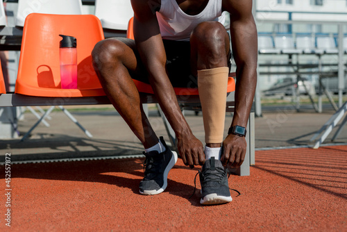 An African American male track and field athlete is sitting on a bench wearing a sports stocking on his left leg before the race