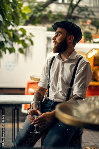 Young bearded man in a cafe on the street with a glass of wine. Romantic guy in a white shirt cap and suspenders in the city. Peaky Blinders. old fassion retro.