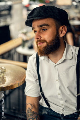 Young bearded man in a cafe on the street with a glass of wine. Romantic guy in a white shirt cap and suspenders in the city. Peaky Blinders. old fassion retro.