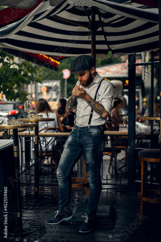 Young bearded man in a cafe on the street smokes a cigarette. Romantic guy in a white shirt cap and suspenders in the city. Peaky Blinders. old fashionable retro.