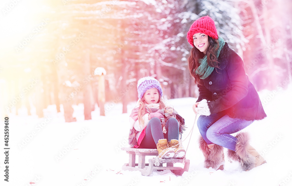 Fototapeta premium A winter fairy tale, a young mother and her daughter ride a sled in the forest. A girl on a sled with gifts on the eve of the new year in the park. 