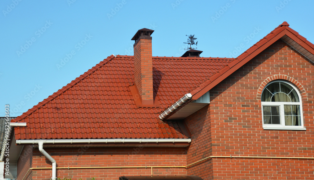 A red metal roofing construction of a red brick house with roof gutters, downpipe, soffit, fascia and box end, an attic window, chimney with roof flashing, a weather vane, and outside gas line.