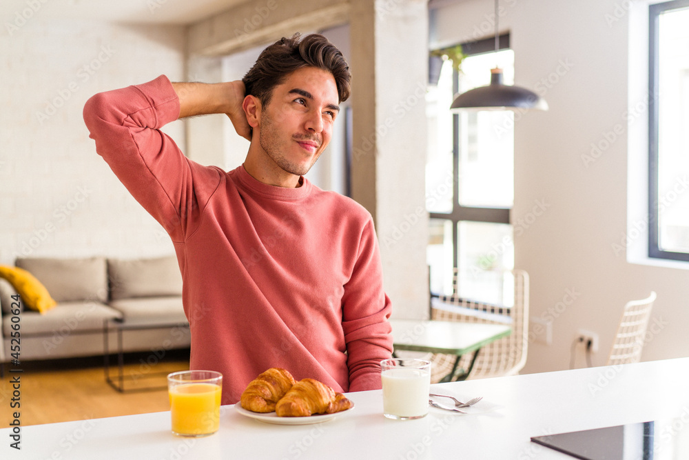 Young mixed race man having breakfast in a kitchen on the morning touching back of head, thinking and making a choice. Young mixed race man having breakfast in a kitchen on the morning touching back of head, thinking and making a choice.