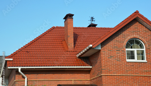 A red metal roofing construction of a red brick house with roof gutters, downpipe, soffit, fascia and box end, an attic window, chimney with roof flashing, a weather vane, and outside gas line.