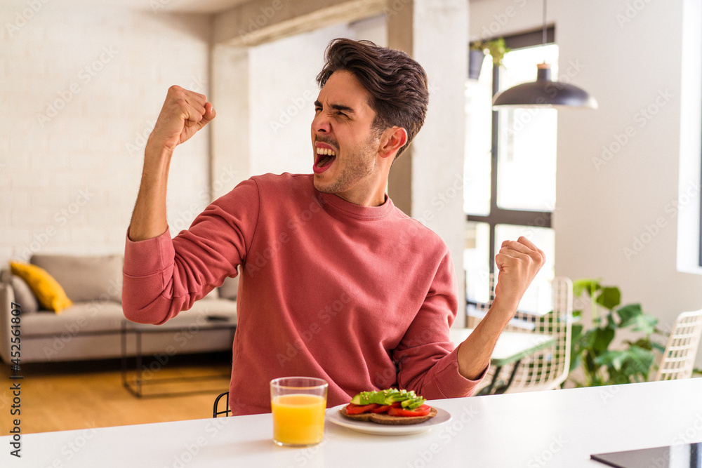 Young mixed race man having breakfast in his kitchen raising fist after a victory, winner concept. Young mixed race man having breakfast in his kitchen raising fist after a victory, winner concept.
