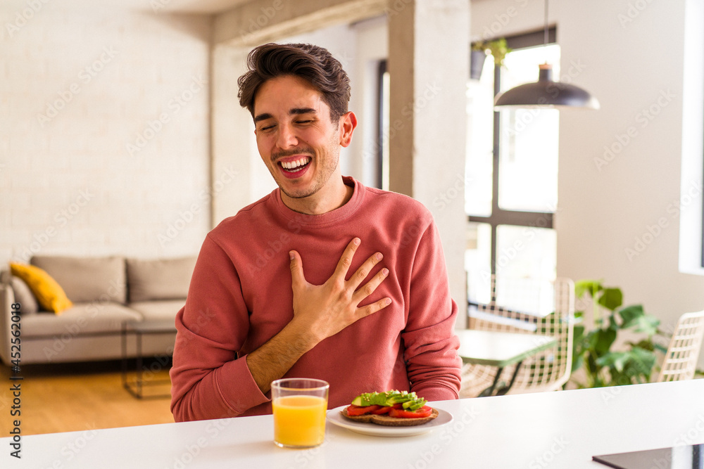 Young mixed race man having breakfast in his kitchen laughs out loudly keeping hand on chest. Young mixed race man having breakfast in his kitchen laughs out loudly keeping hand on chest.