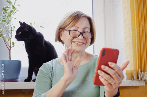 Happy senior woman making video call with smartphone, gesture hi,having pleasant conversation,talking with children grandchildren using wireless internet connection,sitting at home with black pet cat
