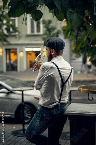 Young bearded tattooed man in a cafe on the street with a glass of wine. A romantic guy in a white cap and suspenders in the city. Peaky Blinders. old fashionable retro.