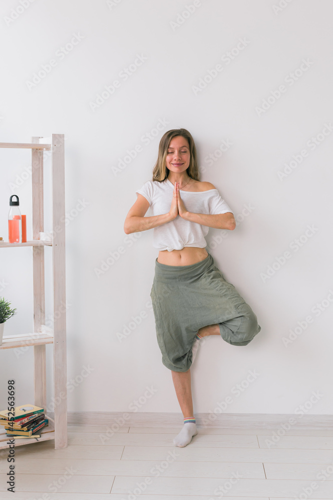 Woman practise yoga performs. Tree pose standing on mat inside of cozy room with plants and greenery. Healthy lifestyle concept