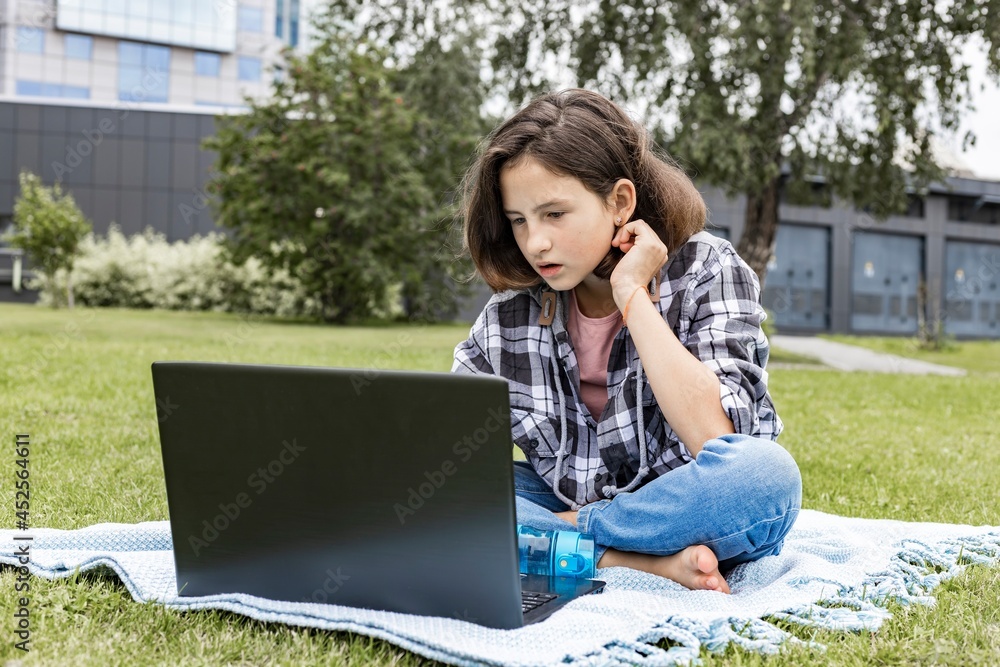 A schoolgirl watches an Internet class and studies remotely. The girl uses wireless internet technologies. A pretty young teenage girl sitting on the grass and working on a laptop. A schoolgirl watches an Internet class and studies remotely. The girl uses wireless internet technologies. A pretty young teenage girl sitting on the grass and working on a laptop.