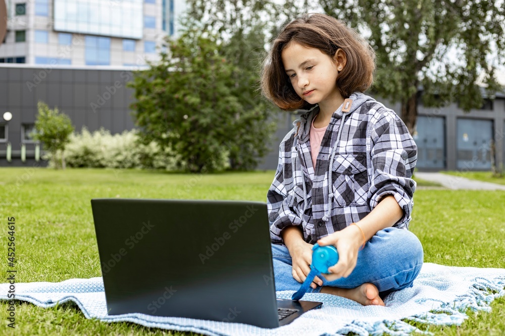 A cute teenage girl sitting on the grass and working on a laptop. A schoolgirl watches an Internet class and studies remotely. The girl uses wireless internet technologies. A cute teenage girl sitting on the grass and working on a laptop. A schoolgirl watches an Internet class and studies remotely. The girl uses wireless internet technologies.