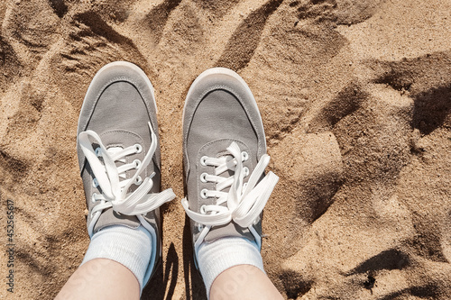 Legs in gray sneakers on beach sand. Summer vacation concept by the sea. Lifestyle travel. Pov view. Copy space.