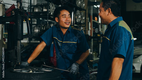 Two professional car mechanic using paperwork makes the oil and engine check to the car on lifted automobile at repair service station at night. Skillful Asian guy in uniform fixing car. Car service.