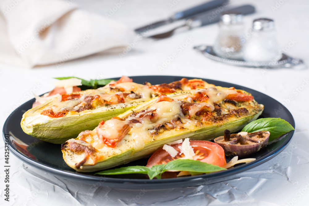 Ready-to-eat baked zucchini halves filled with cheese and tomato and basil leaves on a black plate on a white table. Vegetable menu, healthy food. Close-up Ready-to-eat baked zucchini halves filled with cheese and tomato and basil leaves on a black plate on a white table. Vegetable menu, healthy food. Close-up