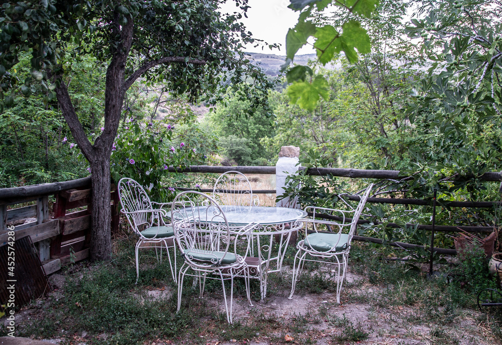 table and four white wrought-iron chairs on a terrace overlooking the countryside with many trees and vegetation next to a thicket with lilac flowers