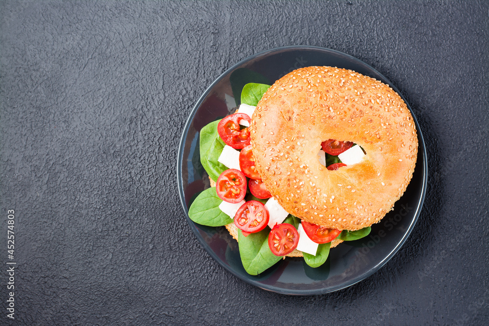 Appetizing ready-to-eat bagels stuffed with tomatoes, feta and spinach leaves on a plate on a black background. Light healthy snack. Top view. Copy space Appetizing ready-to-eat bagels stuffed with tomatoes, feta and spinach leaves on a plate on a black background. Light healthy snack. Top view. Copy space