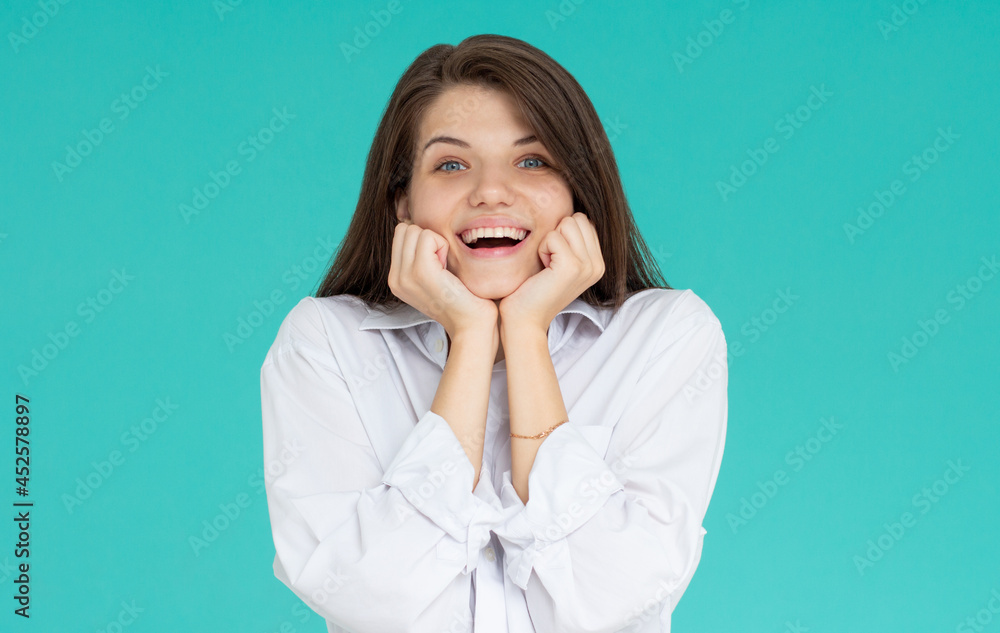Indoor portrait of young attractive European girl isolated on blue background looking straight at camera propping up her chin with fists feeling great joy, smiling and laughing at joke Indoor portrait of young attractive European girl isolated on blue background looking straight at camera propping up her chin with fists feeling great joy, smiling and laughing at joke
