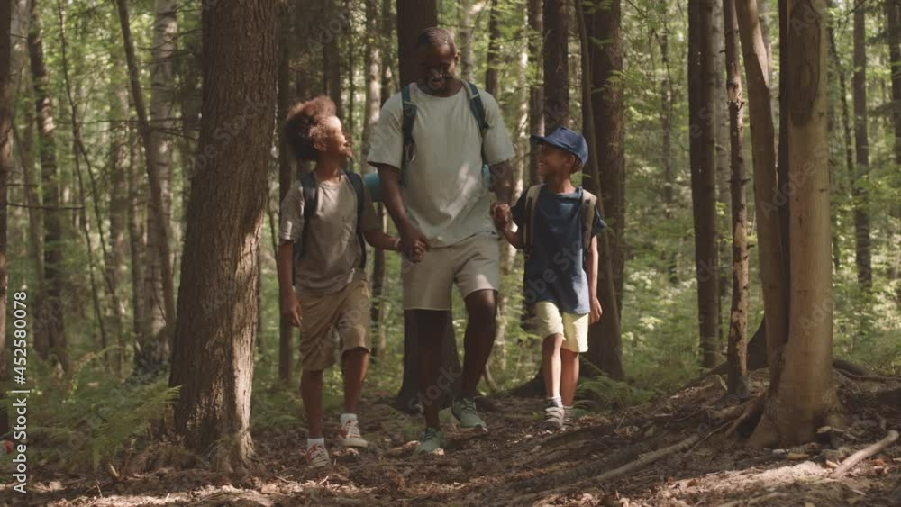 Full-length slowmo shot of African American man holding two little sons by hands while walking towards camera among trees along beautiful summer forest during camping trip Full-length slowmo shot of African American man holding two little sons by hands while walking towards camera among trees along beautiful summer forest during camping trip