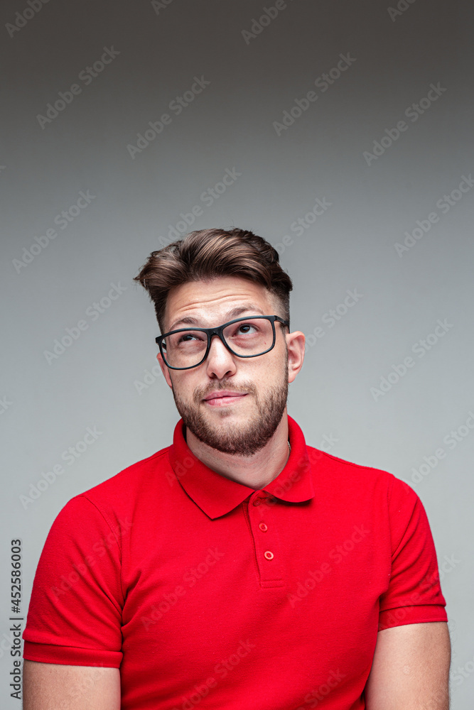 Pensive modern young man in glasses looking up on empty space wrinkling, studio portrait on gray background Pensive modern young man in glasses looking up on empty space wrinkling, studio portrait on gray background